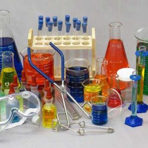 انواع ظروف رنگارنگ آزمایشگاهی، لوله‌های آزمایش، منجوق، فلاسک، وسایل آزمایشگاهی و عینک ایمنی که روی میز چیده شده‌اند، حاوی مواد شیمیایی به رنگ‌های قرمز، آبی، سبز، زرد و نارنجی.